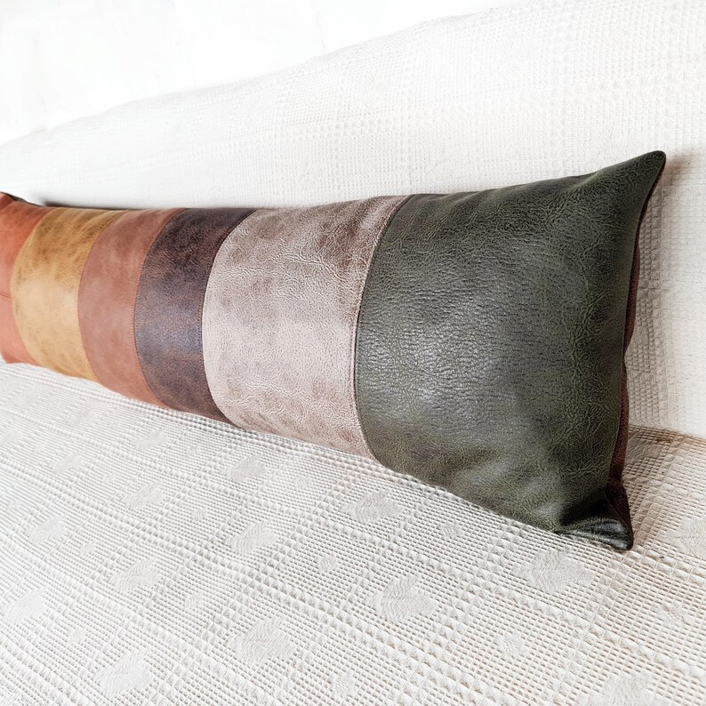 May include: A long rectangular lumbar pillow featuring a color-blocked design with vertical panels in shades of orange, tan, brown, dark brown, light gray, and dark olive green, all crafted from a textured material resembling leather.