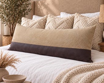 May include: A long rectangular lumbar pillow featuring a two-tone design with a textured tan upper section and a smooth dark brown lower band. The pillow is positioned horizontally across a bed with white bedding and three square textured pillows.