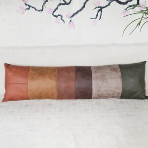 May include: A long, rectangular lumbar pillow featuring six vertical panels of faux leather in varying shades of brown, tan, gray, and dark green. The material has a textured, matte finish with visible stitching between each distinct color block.