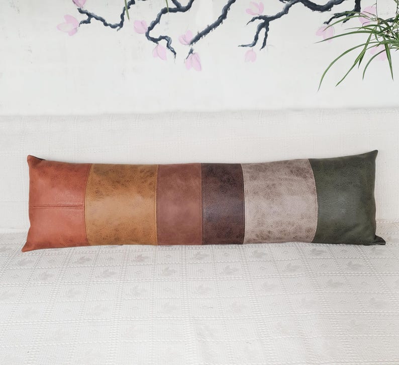 May include: A long, rectangular lumbar pillow featuring six vertical panels of faux leather in varying shades of brown, tan, gray, and dark green. The material has a textured, matte finish with visible stitching between each distinct color block.