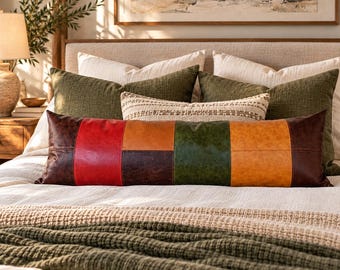 May include: A long rectangular lumbar pillow featuring a patchwork design of faux leather panels in red, dark brown, tan, and forest green. The pillow is positioned on a bed with white bedding and olive green textured throw pillows.