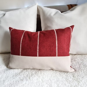 May include: A red and white striped throw pillow with a white faux leather bottom. The pillow is on a white fluffy surface.