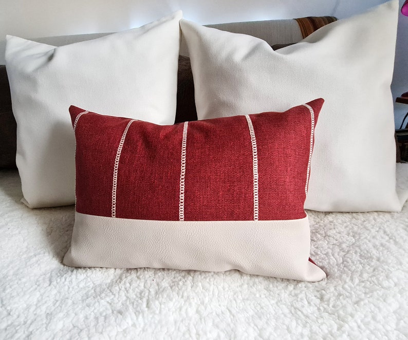 May include: A red and white striped throw pillow with a white faux leather bottom. The pillow is on a white fluffy surface.