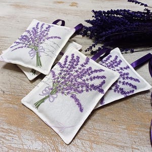 Op de afbeelding: Vier witte stoffen lavendelzakjes met bedrukte paarse lavendelbloemen. De zakjes zijn vastgebonden met een lint en zijn gerangschikt op een houten oppervlak.