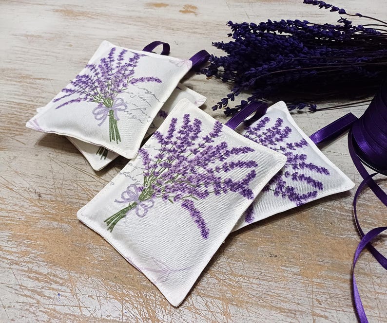 Op de afbeelding: Vier witte stoffen lavendelzakjes met bedrukte paarse lavendelbloemen. De zakjes zijn vastgebonden met een lint en zijn gerangschikt op een houten oppervlak.