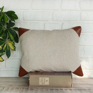 May include: A beige linen pillow with brown leather trim. The pillow is sitting on a book with the title "The Complete Sherlock Holmes" on the spine.