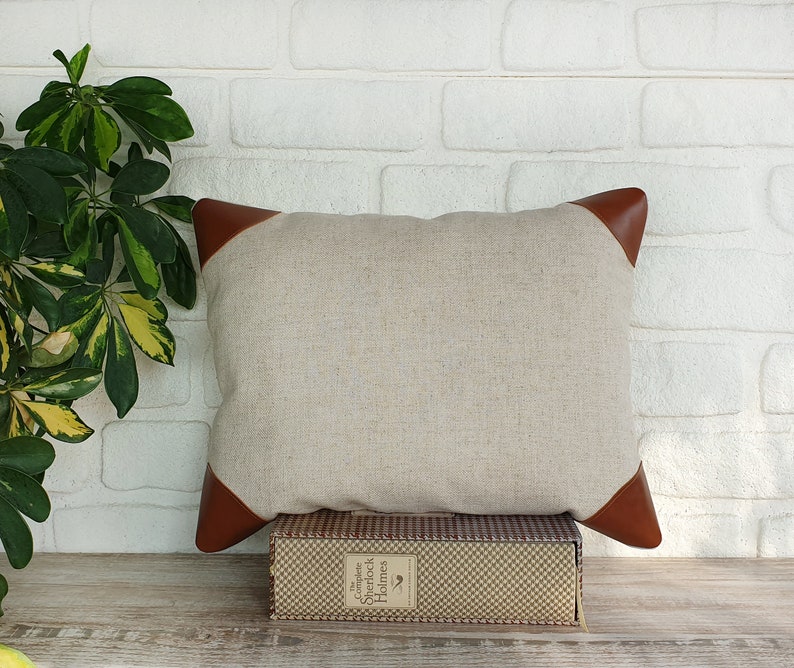May include: A beige linen pillow with brown leather trim. The pillow is sitting on a book with the title "The Complete Sherlock Holmes" on the spine.