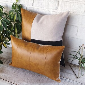 Puede incluir: Dos almohadas decorativas, una con una funda de piel sint&eacute;tica marr&oacute;n y la otra con una funda de lino beige con una franja negra. Las almohadas est&aacute;n dispuestas sobre una superficie de madera.