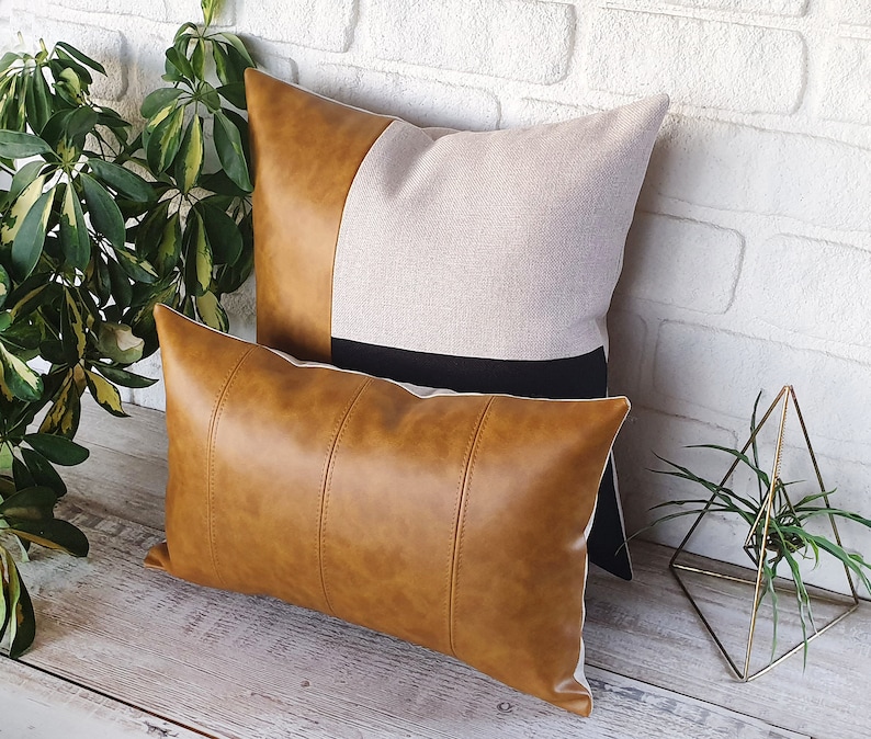 Puede incluir: Dos almohadas decorativas, una con una funda de piel sint&eacute;tica marr&oacute;n y la otra con una funda de lino beige con una franja negra. Las almohadas est&aacute;n dispuestas sobre una superficie de madera.