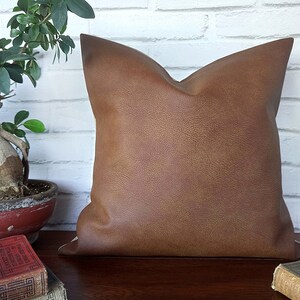 May include: A brown faux leather pillow with a subtle textured surface. The pillow is square and has a simple design.