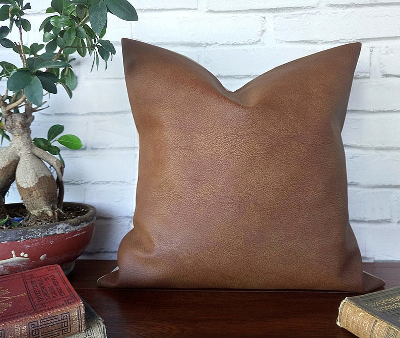 May include: A brown faux leather pillow with a subtle textured surface. The pillow is square and has a simple design.