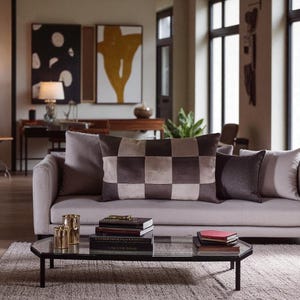 May include: A living room scene featuring a light gray sofa with decorative pillows. A checkered brown and beige pillow is the focal point. A glass-topped coffee table holds books and gold-colored candle holders. Artwork hangs on the wall.