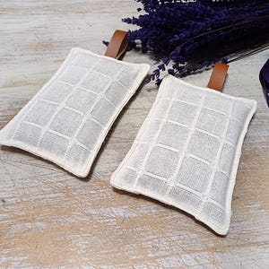 May include: Two white linen sachets with brown leather ties. The sachets have a grid pattern and are filled with dried lavender.