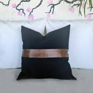Puede incluir: Almohada negra con una banda de cuero marr&oacute;n en el centro. La almohada est&aacute; en un sof&aacute; blanco.