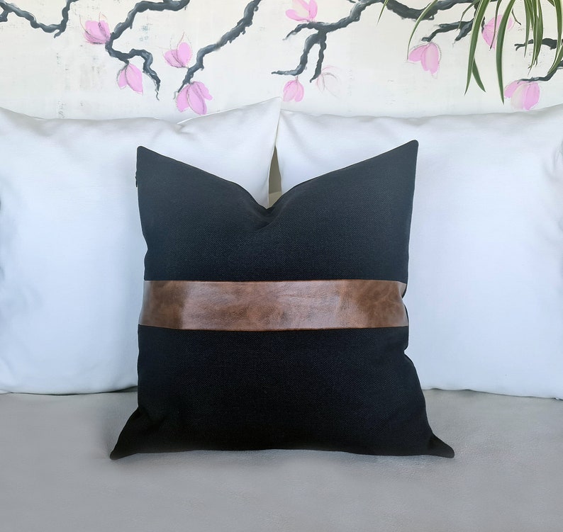 Puede incluir: Almohada negra con una banda de cuero marr&oacute;n en el centro. La almohada est&aacute; en un sof&aacute; blanco.