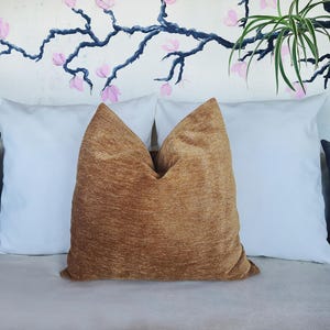 Op de afbeelding: Een decoratief sierkussen met een getextureerde, goudbruine hoes staat tussen twee witte kussens. De kussens liggen op een lichtgrijze ondergrond, met een bloemenmuurschildering op de achtergrond.