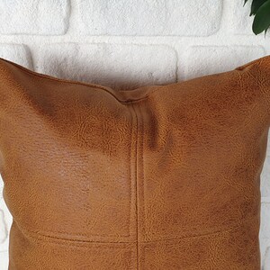 May include: A brown faux leather pillow with a stitched design. The pillow is against a white brick wall.