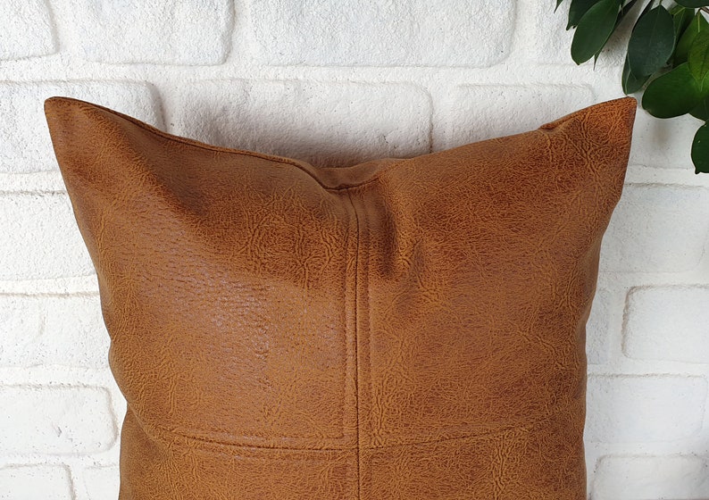 May include: A brown faux leather pillow with a stitched design. The pillow is against a white brick wall.