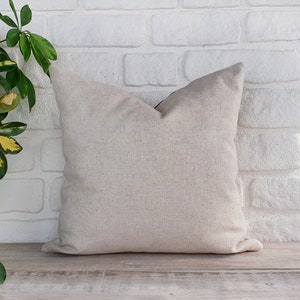 May include: A plain, off-white throw pillow with a subtle texture. The pillow is sitting on a light wood surface in front of a white brick wall.