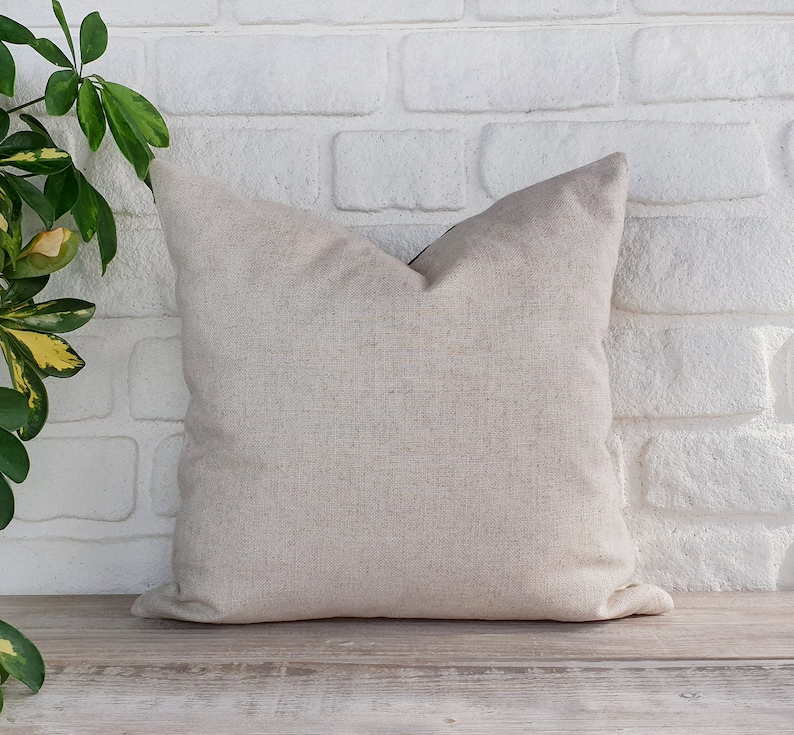 May include: A plain, off-white throw pillow with a subtle texture. The pillow is sitting on a light wood surface in front of a white brick wall.