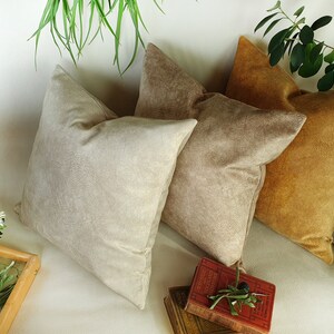 May include: Three decorative throw pillows in beige, light brown, and mustard yellow. The pillows are made of a soft, textured fabric and are arranged on a white surface.