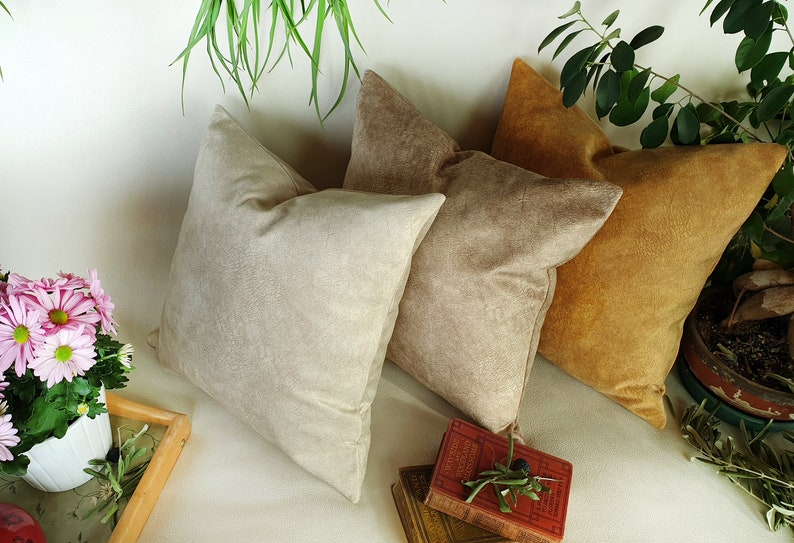 May include: Three decorative throw pillows in beige, light brown, and mustard yellow. The pillows are made of a soft, textured fabric and are arranged on a white surface.