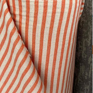 May include: Orange and white striped fabric with a subtle texture. The fabric is rolled up and the stripes are vertical.