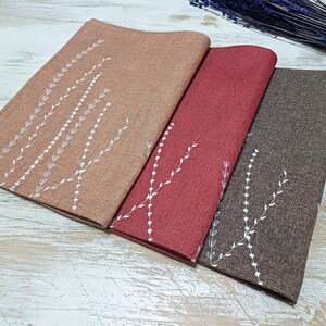 May include: Three placemats with a white embroidered design of branches and leaves on a brown, red, and beige background.