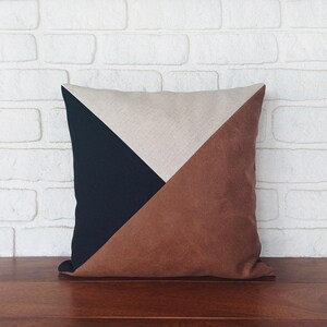 May include: Square decorative pillow with a geometric design. The pillow features a color block pattern with sections of navy blue, tan, and brown. The pillow is placed on a wooden surface against a white brick wall.