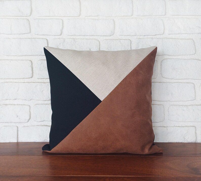 May include: Square decorative pillow with a geometric design. The pillow features a color block pattern with sections of navy blue, tan, and brown. The pillow is placed on a wooden surface against a white brick wall.