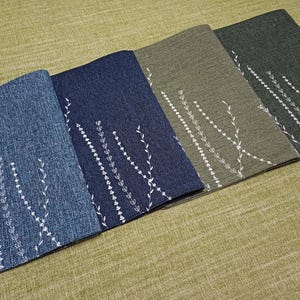 May include: Four placemats with a white floral design on a textured fabric background. The placemats are in shades of blue, green, and brown.