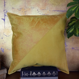 May include: A square cushion with a two-tone design. One half is textured, golden-yellow faux leather, the other a solid, mustard-yellow fabric. The cushion is placed on a stack of books against a gold-toned background.