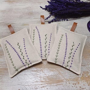 May include: Three small, rectangular linen sachets with lavender embroidery. The sachets are beige with purple and green stitching. The sachets have brown leather ties.
