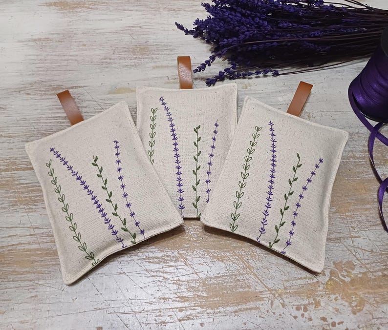 May include: Three small, rectangular linen sachets with lavender embroidery. The sachets are beige with purple and green stitching. The sachets have brown leather ties.