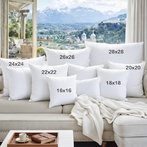 May include: A collection of white square and rectangular throw pillows of various sizes, including 16x16, 18x18, 20x20, 22x22, 24x24, 26x26, and 28x28 inches, arranged on a beige sofa. A white blanket is draped over the sofa.