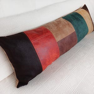 May include: A long rectangular throw pillow featuring a patchwork design of faux leather panels in black, red, tan, dark brown, and dark green. The material has a textured, distressed finish across all the coloured sections of the pillow.