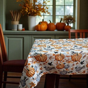 Könnte beinhalten: Ein weißes Tischtuch mit einem Kürbis- und Blättermuster. Die Kürbisse sind orange, grau und weiß, mit schwarzen Blättern und orangefarbenen floralen Akzenten. Der Tisch ist für ein Herbstfest gedeckt.
