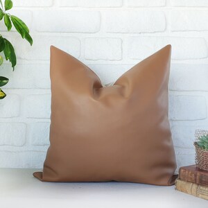 May include: A brown leather pillow with a square shape and a smooth texture. The pillow is sitting on a white brick wall surface.