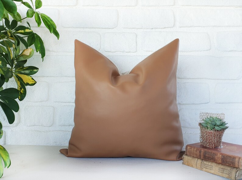 May include: A brown leather pillow with a square shape and a smooth texture. The pillow is sitting on a white brick wall surface.