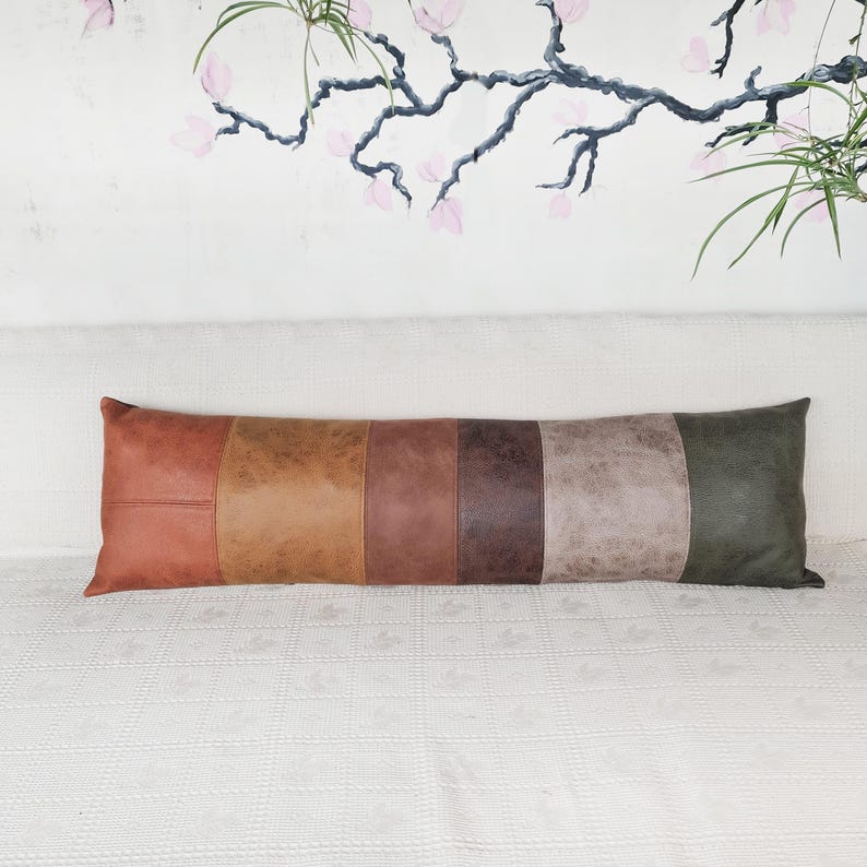 May include: A long, rectangular lumbar pillow featuring a color-blocked design made of six vertical panels in varying shades of leather-like material, including rust, tan, brown, dark chocolate, taupe, and olive green with a smooth texture.