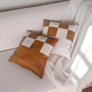 May include: Two decorative throw pillows with a checkered pattern. The pillows are made of a brown faux leather and white fabric. The pillows are on a white couch.