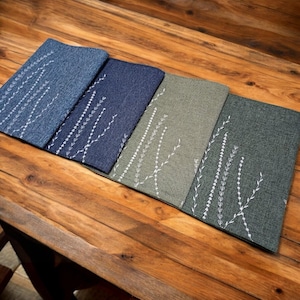 Könnte beinhalten: Vier Platzsets mit einem weißen, gestickten Blumenmuster. Die Platzsets sind in Blautönen und Grün.