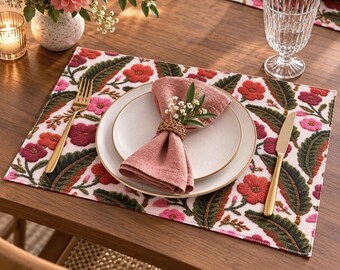 May include: A rectangular placemat featuring an embroidered floral pattern in shades of pink, red, and dark green on a white background. A white ceramic plate holds a folded dusty pink linen napkin secured with a woven ring and a small floral sprig.
