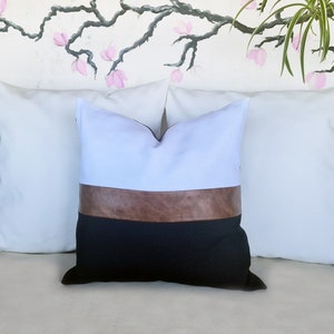 Puede incluir: Una almohada decorativa blanca, marr&oacute;n y negra con un acento de cuero. La almohada est&aacute; sentada en un sof&aacute; blanco.
