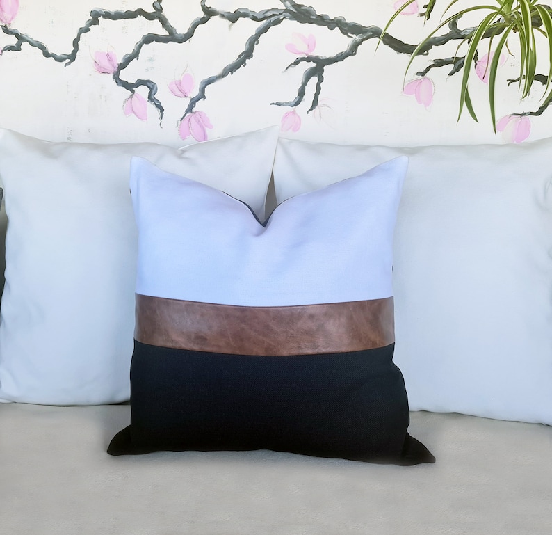 Puede incluir: Una almohada decorativa blanca, marr&oacute;n y negra con un acento de cuero. La almohada est&aacute; sentada en un sof&aacute; blanco.
