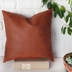 May include: A brown faux leather pillow with a textured surface. The pillow is sitting on a book with a white and brown checkered pattern.