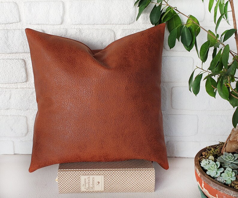 May include: A brown faux leather pillow with a textured surface. The pillow is sitting on a book with a white and brown checkered pattern.