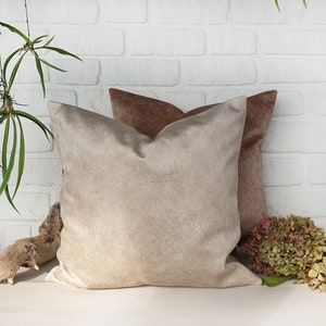 May include: Two decorative throw pillows, one beige and one brown, with a soft, textured fabric. The pillows are arranged on a white brick wall background.