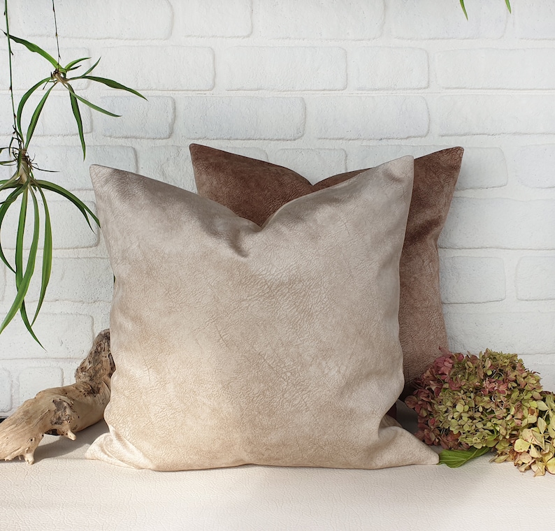 May include: Two decorative throw pillows, one beige and one brown, with a soft, textured fabric. The pillows are arranged on a white brick wall background.