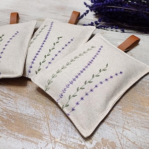 May include: Three beige linen sachets with lavender embroidery. The sachets are filled with dried lavender and are perfect for adding a touch of fragrance to any room.